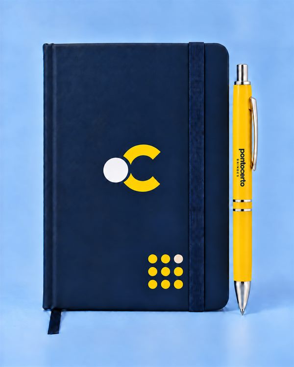 Caderneta de capa dura emborrachada azul-marinho com elástico vertical e o símbolo da Ponto Certo Brindes na capa, ao lado de uma caneta amarela personalizada.